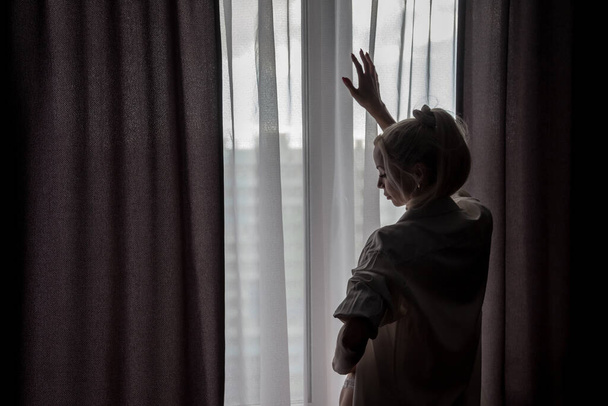 Silhouette of girl in white men shirt at window. Portrait of nice female. Home comfort. Slavic woman is standing and touches curtain of tulle. Emotions and relax. Concept image - Φωτογραφία, εικόνα