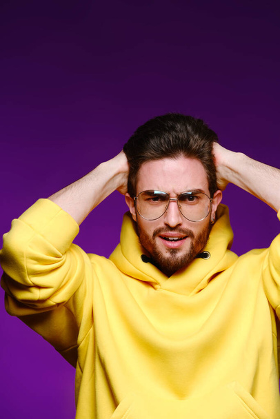 A young man of 25-30 years in glasses and a yellow sweatshirt emotionally poses holding his head on a purple background. - Zdjęcie, obraz