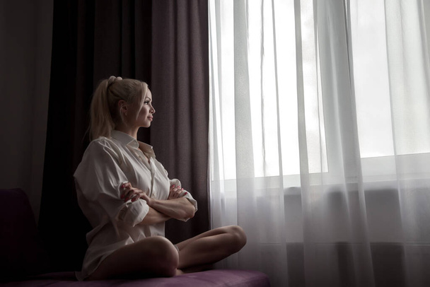 Girl on sofa at window of house waiting at sunlight from window. Portrait of nice woman. Home comfort and coziness. Slavic female in morning in room. Emotions and relax. Concept of home relaxation - Foto, Imagen