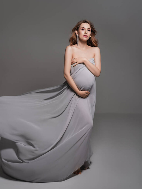 Těhotná žena zabalená v závoji drží břicho s pažem.Módní portrét šťastné těhotné ženy.Těhotenství, mateřství, příprava a očekávání koncepce.Těhotná žena v černém těle. Glamour a módní mateřství portrét na šedém pozadí - Fotografie, Obrázek