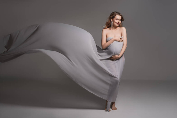 Pregnant woman wrapped in veil holding her belly with arms.Fashion portrait of happy pregnant woman.Pregnancy, maternity, preparation and expectation concept.Pregnant woman in black body. Glamour and fashion motherhood portrait on gray background - Foto, Imagen