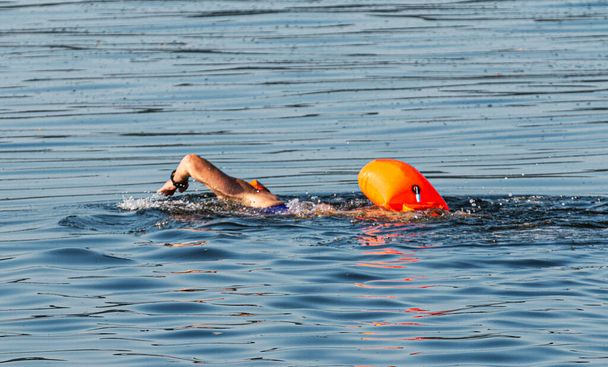 Yüzen bir adam körfezde turuncu bir yüzgeçle sürünüyor ve arkasından triatlon antrenmanı için yüzüyor.. - Fotoğraf, Görsel