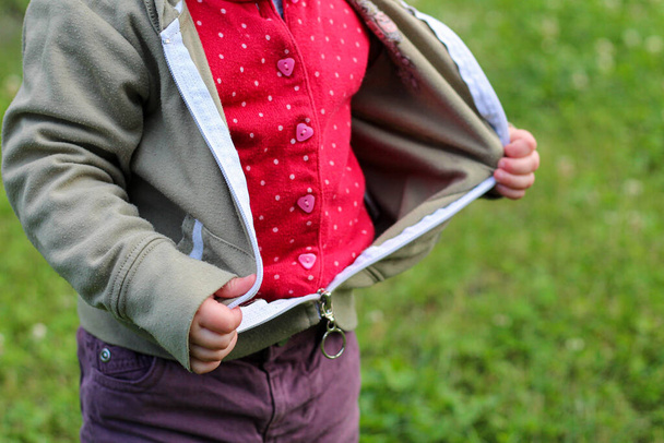 Los mangos de los niños pequeños desabrochan la cremallera de una chaqueta sobre un fondo de hierba verde borrosa
 - Foto, Imagen