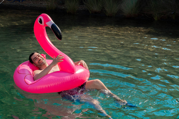 Leuke en grappige man genieten op zijn opgeblazen roze flamingo in zonnige dag.  - Foto, afbeelding