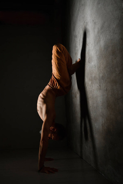 Aged male with naked torso, in brown pants is practicing yoga. Standing on hands, leaning against wall by legs being in lotus pose - Fotó, kép