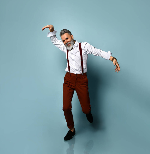 Elderly man in white shirt, brown pants, suspenders, black loafers. Smiling, running, waving his hands, posing on blue background - Фото, изображение