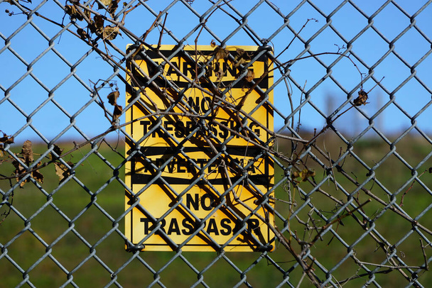 Advertencia: No hay señal de allanamiento en inglés y español parcialmente oculta detrás de una valla ciclónica, con ramas entrelazadas
. - Foto, imagen