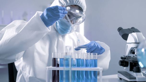 Επιστήμονας στη σουίτα PPE κάνει κάποια έρευνα ελέγχοντας ένα υγρό σε ένα δοκιμαστικό σωλήνα στο εργαστήριο. - Φωτογραφία, εικόνα