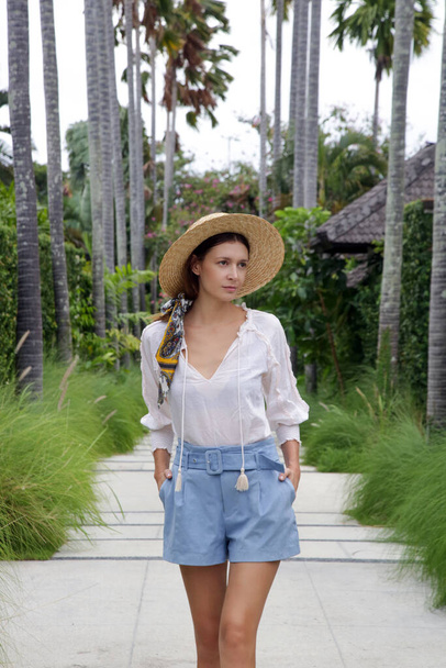 Fashionable tourist woman wearing straw hat walking on the lane between tall palm trees. Tropical summer vacation concept. - Photo, Image
