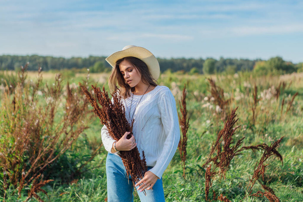 Happy beautiful brunette teenage girl dressed in a white sweater, jeans and cowboy straw hat standing in a field holding dry bouquet - Photo, Image