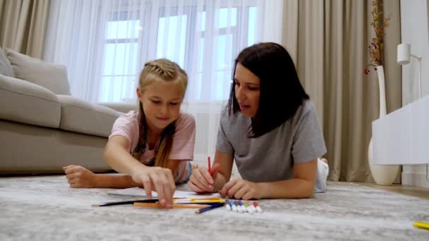 Smiling young mom and cute preteen daughter lying on warm floor at home drawing in album together, happy parent or nanny have fun spending time painting with colorful pencils, slow motion - Footage, Video