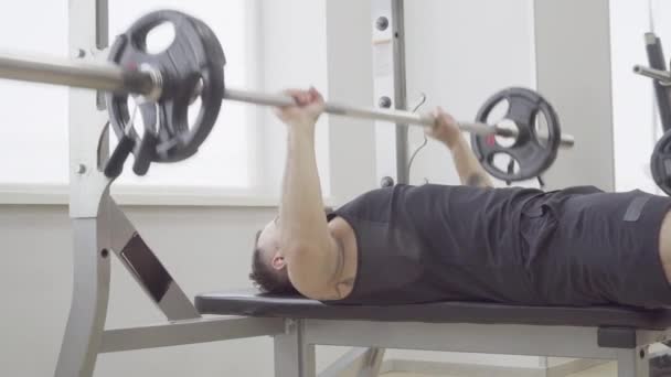 Strong Caucasian sportsman lying on coach and raising barbell. Side view portrait of athletic confident Caucasian man training in gym. Strength, masculinity, sport, healthy lifestyle. - Filmagem, Vídeo