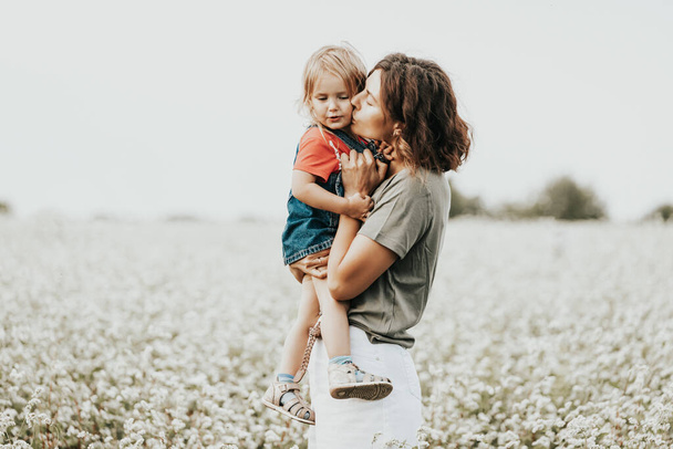 Ein romantisches Porträt einer jungen Frau und ihrer Tochter mit lockigem Haar spielen und umarmen sich auf einem Feld mit üppigen Blumen. Kreatives Porträt einer Familie in der Natur. Freundschaft zwischen Mutter und Tochter - Foto, Bild