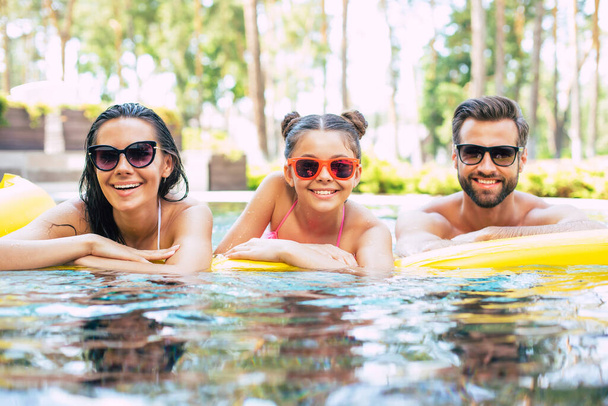Handsome smiling man and cute excited woman with them little lovely daughter are having fun in summer swim pool while resting in hotel. - Photo, image