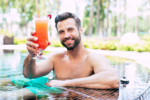 Excited handsome modern muscular man is resting in summer pool and drinks refreshing cocktail and looks on camera. - 写真・画像