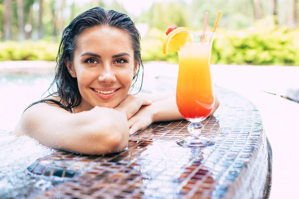 Beautiful excited smiling woman is relaxing in summer swim pool and drinks refreshing cocktail. - Foto, Imagem