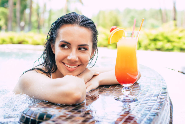 Beautiful excited smiling woman is relaxing in summer swim pool and drinks refreshing cocktail. - Foto, Imagen