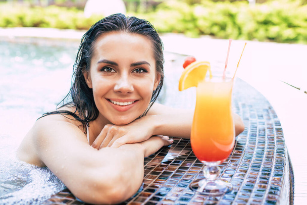 Beautiful excited smiling woman is relaxing in summer swim pool and drinks refreshing cocktail. - Photo, image