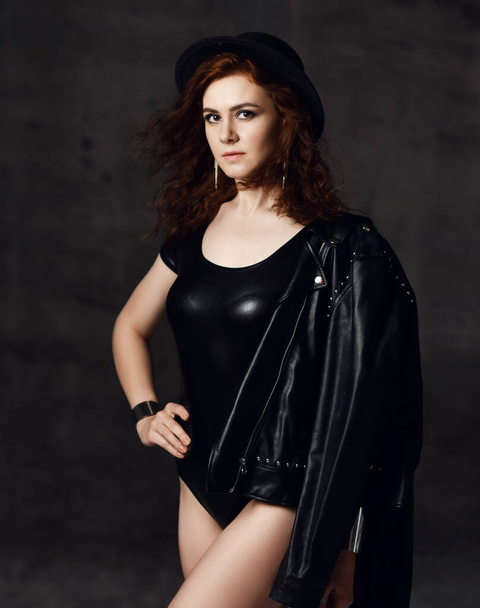 Young red haired curly woman in body underwear, leather jacket, hat and accessories stands with her hand on her waist - Foto, Imagem
