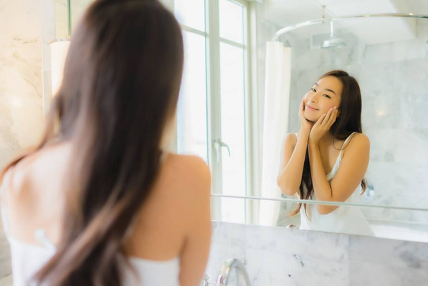 Portré gyönyörű fiatal ázsiai nő ellenőrizze, és töltsük fel az arcát a tükör a fürdőszoba belsejében - Fotó, kép