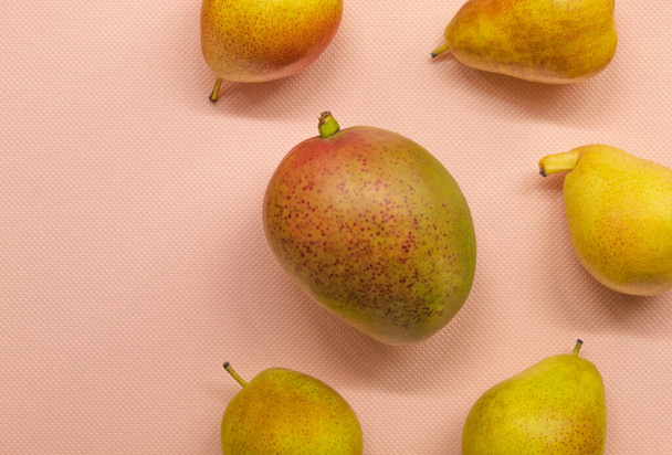 In the horizontal picture on a pink background are fresh fruit. In the center is a small mango, around it yellow pears. Fruits are ready to eat, they are good for health - Photo, Image