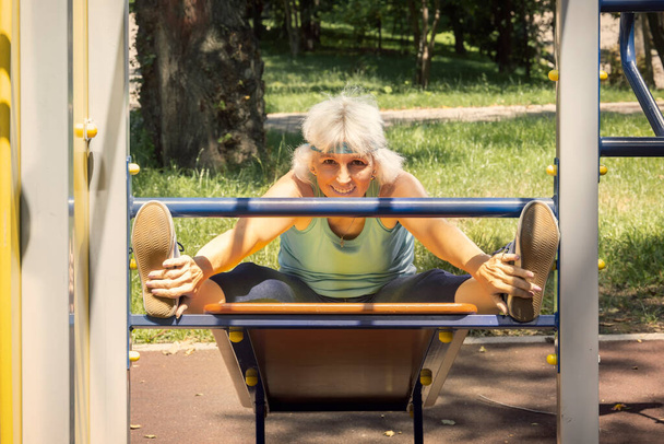 Mulher idosa magra com cabelo grisalho conduz aulas de fitness individuais em um simulador em um parque da cidade - Foto, Imagem