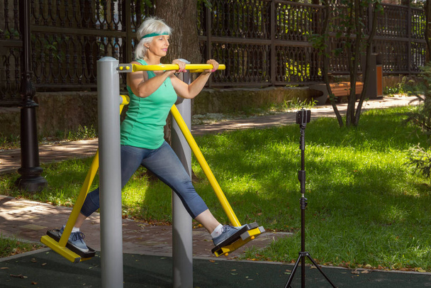 Štíhlá starší žena s šedivými vlasy provádí virtuální trénink pomocí mobilního telefonu na simulátoru v městském parku během jednotlivých fitness tříd - Fotografie, Obrázek