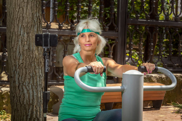 Snella donna anziana con i capelli grigi conduce un allenamento virtuale utilizzando un telefono cellulare su un simulatore in un parco cittadino durante le lezioni di fitness individuali. Primo piano - Foto, immagini