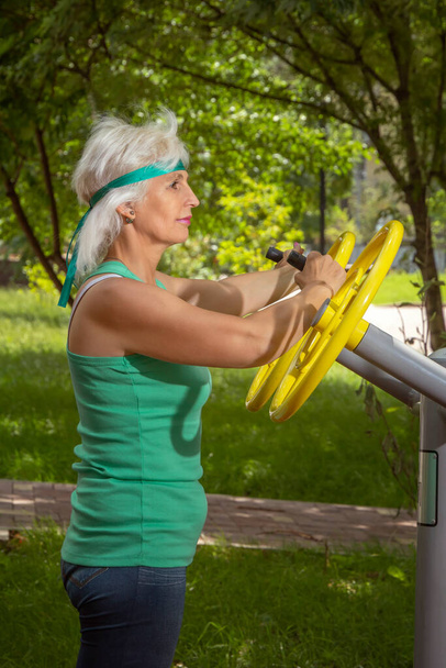 Mujer de edad avanzada delgada con el pelo gris conduce clases individuales de fitness en un simulador en un parque de la ciudad - Foto, imagen