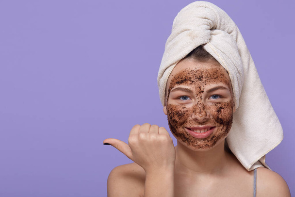 Beautiful relaxed smiling female points thumb finger on free space, applies natural chocolate mask on her face, reducess dryness, shows place for your advertising content. - Valokuva, kuva