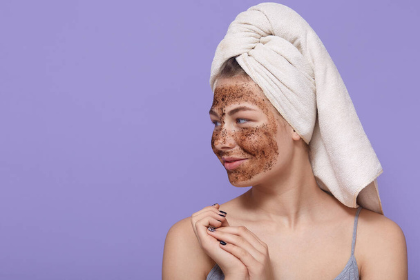 Portrait of female model applies chocolate mask on face, has positive expression, looks aside, wears towel on head after bath, poses over lilac wall with, uses beauty product for skin care. - Фото, изображение