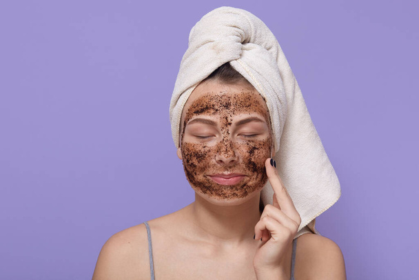 Portrait of young female applies homemade facial clay mask, has white towel wrapped around head, keeping eyes closed, stands with finger on cheek against lilac background. Beauty treatment at home. - Foto, imagen
