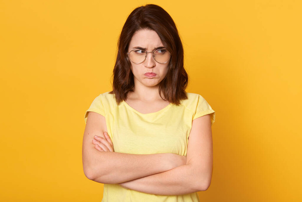 Offended woman wearing yellow t shirt, female with beautiful hair posing with pouty lips, looks upset, standing against yellow wall with folded hands and looking aside. - Фото, зображення