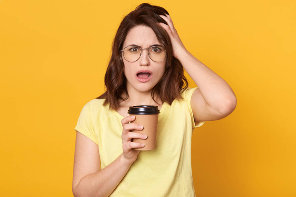 Young beautiful woman wearing glasses and casual t shirt holding take away coffee isolated over yellow background, being in shock, standing with surprise face, opened mouth, keeps hand on head. - Fotografie, Obrázek