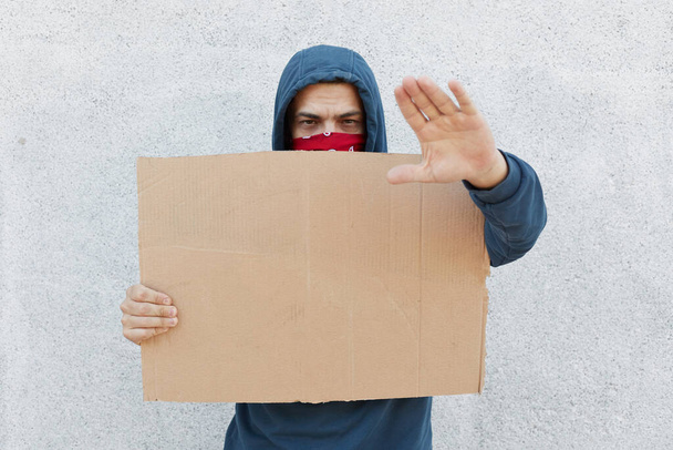Young protesting man in hood and covering his face with bandana stands against white wall holding poster and showing stop sign with palm. Black Lives Matter. - Foto, Bild