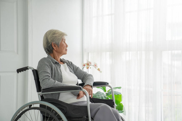 Tekerlekli sandalyede oturan yaşlı bir kadının pencereden dışarı bakışı. Hastane odasındaki yaşlı kadın pencerenin yanında oturup düşünüyor. Yaşlı hasta hastanede üzgün ve yalnız hissediyor.. - Fotoğraf, Görsel