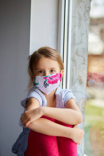 小さな女の子、マスクの子供は窓の上に座って、コロナウイルスの隔離、家庭学校。家にいて。コロナウイルスの隔離の概念。MERS-COV 。新コロナウイルス. - 写真・画像