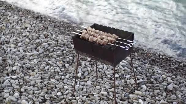 Una pequeña parrilla de senderismo con carbón quemado, brochetas en brochetas, en la playa, en una playa de guijarros
. - Metraje, vídeo