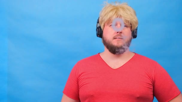 freaky fat man in a wig and a pink T-shirt in his headphones exhales cigarette smoke, smokes a vape against a blue background. - Footage, Video