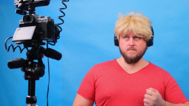 homem gordo estranho em uma peruca e uma camiseta rosa faz um blog de vídeo contra um fundo azul
. - Filmagem, Vídeo