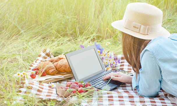 Şapkalı kız parkta bir piknikte dizüstü bilgisayarda çalışıyor. Uzaktan kumanda. Modern hayat, serbest çalışan. Boşluğu kopyala. - Fotoğraf, Görsel