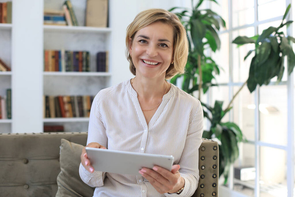 Cheerful middle aged woman sitting on sofa, using computer tablet apps, looking at screen,reading good news in social network, shopping or chatting online - Foto, imagen