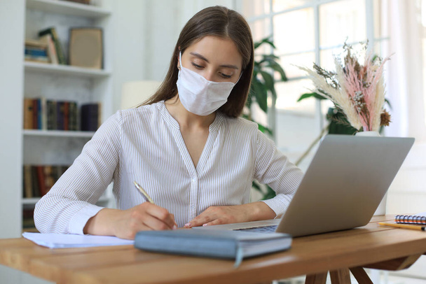 Young businesswoman in a medical protective mask works from home at the laptop during self-isolation and quarantine - Fotoğraf, Görsel