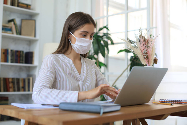 Young businesswoman in a medical protective mask works from home at the laptop during self-isolation and quarantine - Foto, Imagen