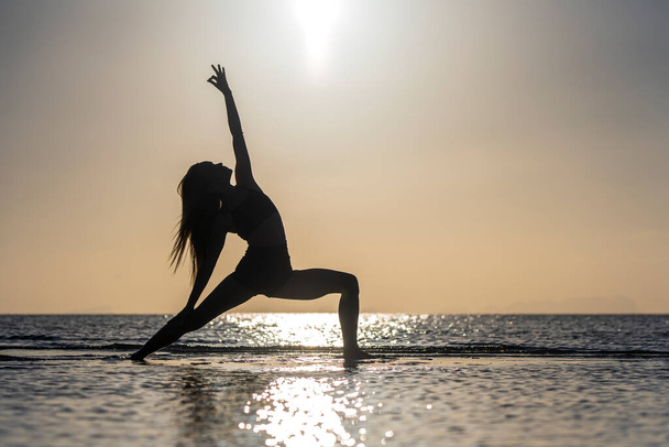 Silhouette of woman standing at yoga pose on the tropical beach during sunset. Caucasian girl practicing yoga near sea water - Fotó, kép