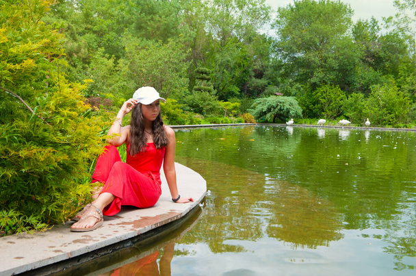 Młoda brunetka pozowała na drewnianym pomoście niedaleko jeziora. Dziewczyna ubrana w czerwony kostium. Jezioro Łabędzie, plener. - Zdjęcie, obraz