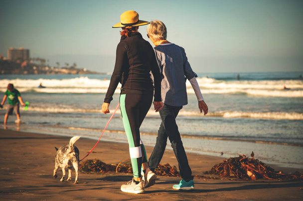 Дружба. Жінки ходять на пляжі, з собакою. Молода жінка і старша жінка ходять разом, в сонячний день, на пляжі
. - Фото, зображення
