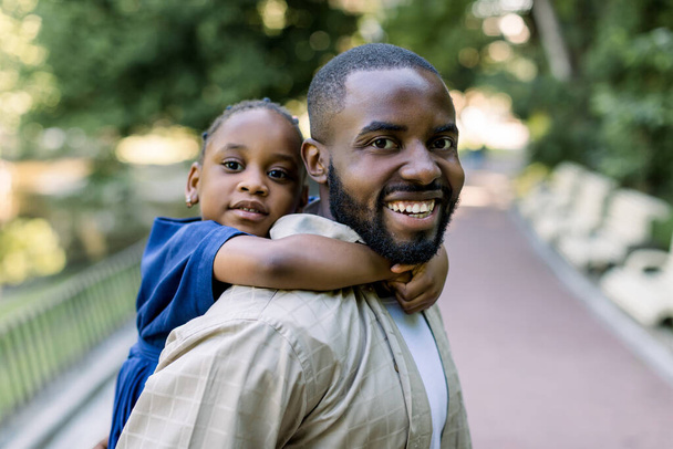 Smiling handsome African American father carrying his cute little daughter on back outdoors in city park. Daddy carrying her daughter on piggyback in the nature. - Φωτογραφία, εικόνα