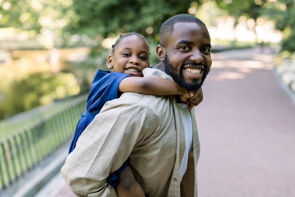 Handsome young dark skinned father and his laughing little daughter, playing in the park. African father piggyback his little daughter outside in nature. Close up portrait - Φωτογραφία, εικόνα