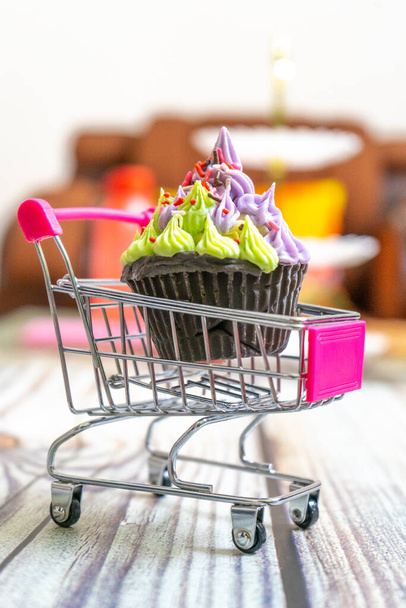 Beautifully iced cupcake with purple frosting and black chocolate base on a shopping cart on a wooden floor with blurred out background - Photo, Image